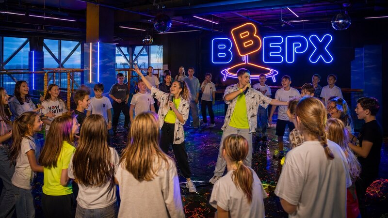 Много подростков и ведущие танцуют под объёмным неоновым логотипом Вверх